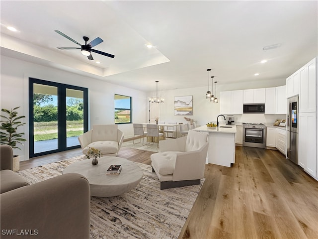 living room with ceiling fan with notable chandelier, light hardwood / wood-style floors, sink, and a tray ceiling