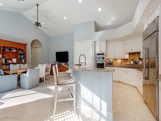 kitchen with high vaulted ceiling, ceiling fan, an island with sink, appliances with stainless steel finishes, and tasteful backsplash