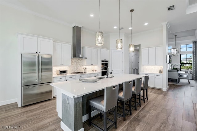 kitchen with wall chimney exhaust hood, stainless steel appliances, a kitchen island with sink, white cabinets, and light hardwood / wood-style floors