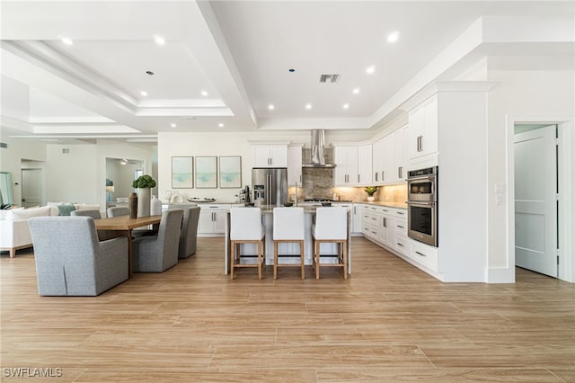 kitchen with a kitchen bar, stainless steel appliances, wall chimney range hood, a center island with sink, and white cabinetry