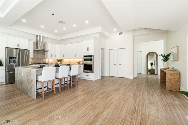 kitchen with a center island with sink, a kitchen breakfast bar, white cabinetry, and wall chimney range hood