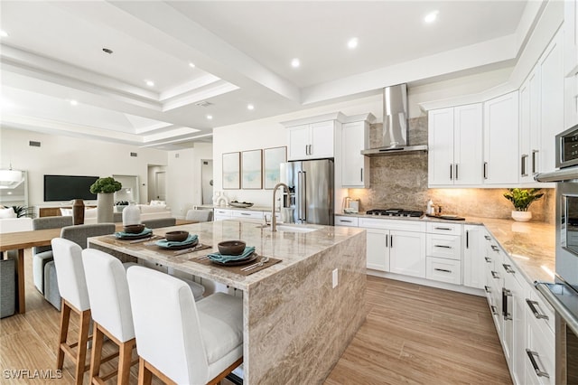 kitchen with sink, stainless steel appliances, wall chimney range hood, light stone counters, and a kitchen island with sink