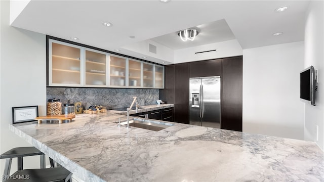 kitchen with backsplash, glass insert cabinets, light stone countertops, stainless steel refrigerator with ice dispenser, and a sink