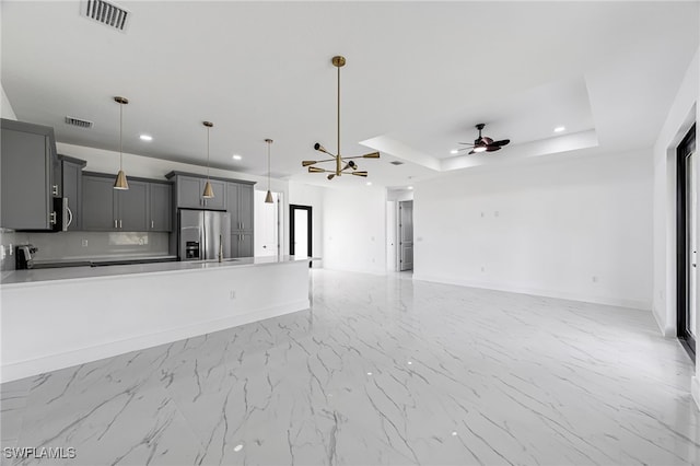 unfurnished living room featuring a tray ceiling, a wealth of natural light, and ceiling fan with notable chandelier