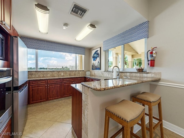 kitchen featuring kitchen peninsula, light stone counters, stainless steel appliances, light tile patterned floors, and a breakfast bar area