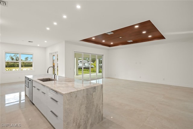 kitchen featuring light stone counters, a tray ceiling, sink, white cabinetry, and an island with sink