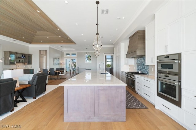 kitchen featuring appliances with stainless steel finishes, light wood-type flooring, custom range hood, a large island with sink, and decorative light fixtures