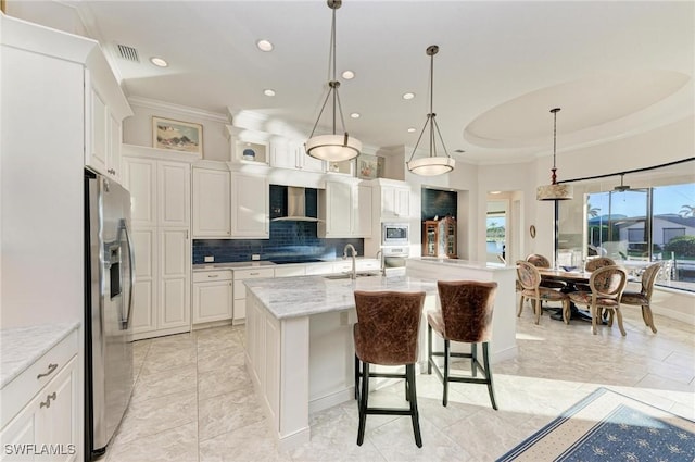 kitchen with wall chimney range hood, white cabinetry, light stone countertops, and a center island with sink