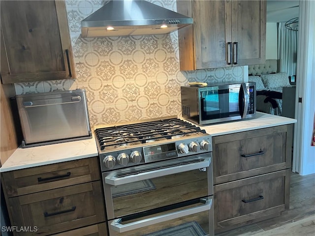 kitchen featuring dark brown cabinetry, wall chimney exhaust hood, light wood-type flooring, and appliances with stainless steel finishes