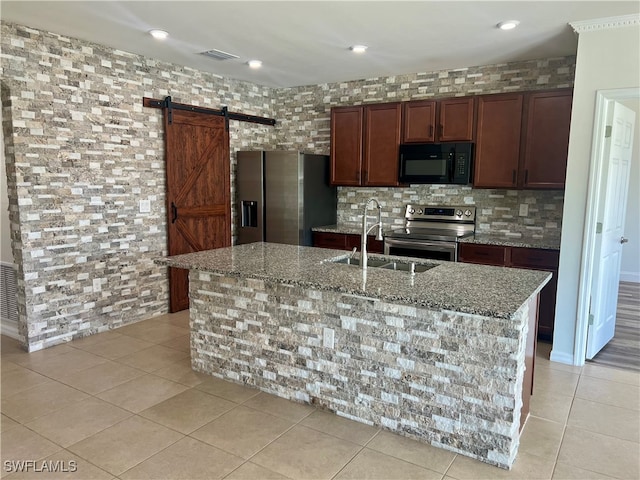 kitchen with sink, a barn door, light stone counters, an island with sink, and appliances with stainless steel finishes