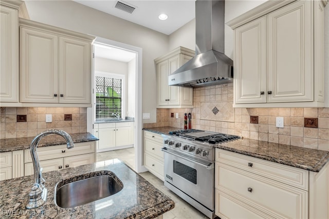 kitchen with dark stone counters, sink, wall chimney range hood, high end stainless steel range, and cream cabinetry