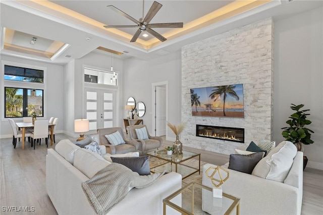 living room with ceiling fan, a stone fireplace, light wood-type flooring, and a tray ceiling