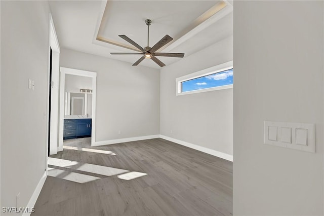 unfurnished bedroom featuring a tray ceiling, ceiling fan, ensuite bath, wood finished floors, and baseboards