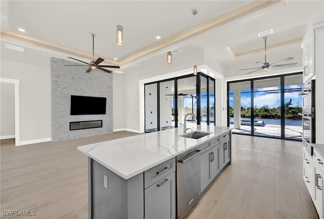 kitchen with stainless steel dishwasher, a tray ceiling, sink, a center island with sink, and white cabinetry