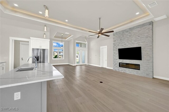 unfurnished living room with ceiling fan, sink, a raised ceiling, a stone fireplace, and light hardwood / wood-style flooring