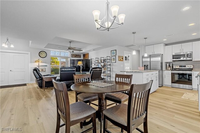 dining room with light wood-type flooring, ceiling fan with notable chandelier, and a tray ceiling