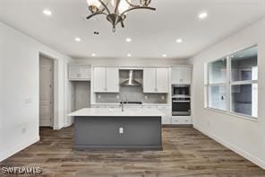 kitchen featuring an inviting chandelier, a center island with sink, wall chimney exhaust hood, dark hardwood / wood-style flooring, and white cabinetry