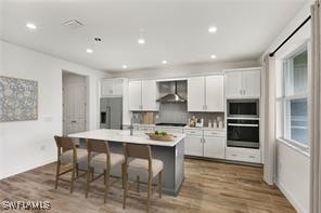 kitchen with a kitchen breakfast bar, stainless steel appliances, a kitchen island with sink, wall chimney range hood, and white cabinetry