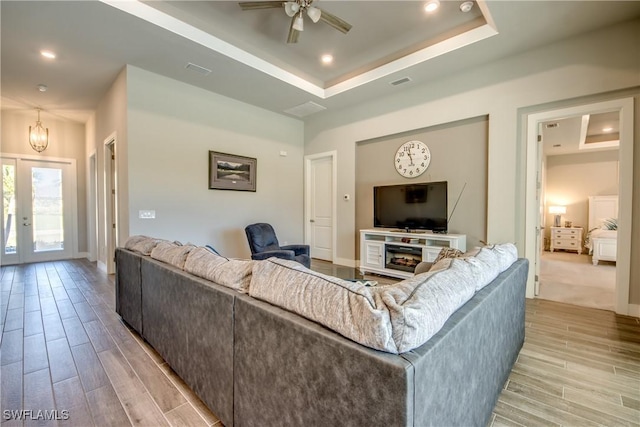 living room featuring ceiling fan with notable chandelier, french doors, light wood-type flooring, and a tray ceiling