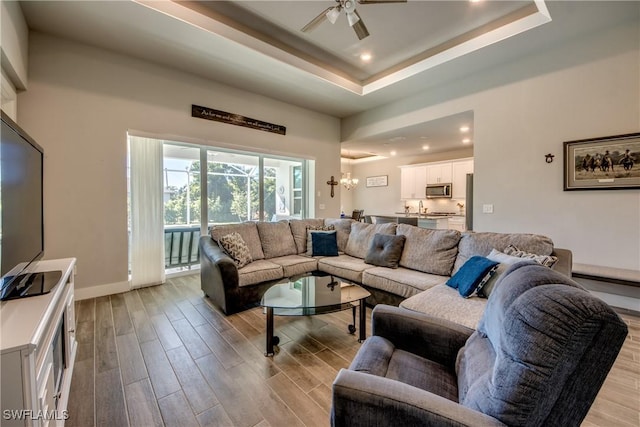 living room featuring ceiling fan with notable chandelier, light wood-type flooring, and a raised ceiling