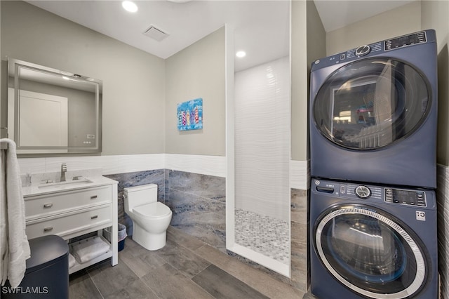 clothes washing area with sink, stacked washing maching and dryer, and tile walls