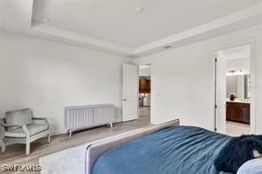 bedroom with a raised ceiling, ensuite bathroom, radiator heating unit, and light hardwood / wood-style floors