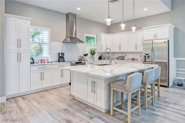 kitchen with white cabinetry, wall chimney range hood, stainless steel appliances, and an island with sink