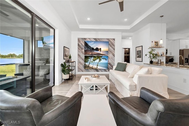 living room with ceiling fan, light wood-type flooring, sink, and a tray ceiling