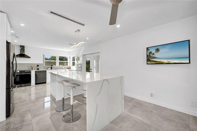 kitchen featuring stainless steel appliances, wall chimney range hood, decorative light fixtures, a center island, and white cabinetry