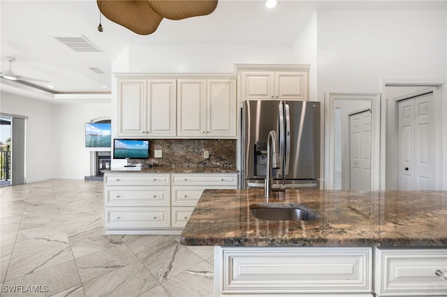 kitchen with ceiling fan, backsplash, stainless steel fridge, dark stone counters, and a center island with sink