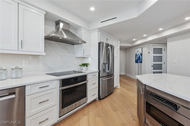 kitchen featuring appliances with stainless steel finishes, wall chimney range hood, light stone countertops, decorative backsplash, and white cabinets