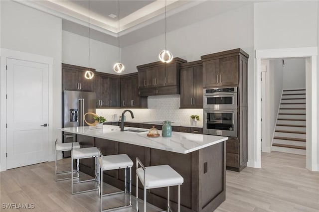 kitchen with stainless steel appliances, a high ceiling, a kitchen island with sink, dark brown cabinets, and light wood-type flooring