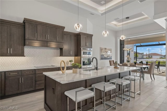 kitchen featuring black electric cooktop, an island with sink, stainless steel double oven, and decorative light fixtures