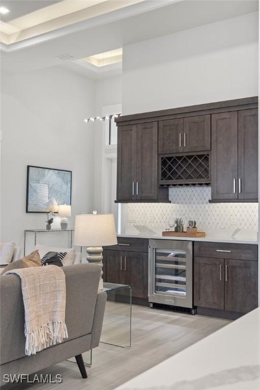 bar featuring backsplash, wine cooler, dark brown cabinetry, and light wood-type flooring