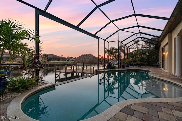 pool at dusk with a patio area, glass enclosure, a water view, and a dock