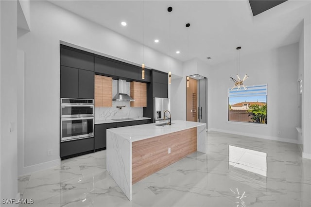 kitchen featuring pendant lighting, a kitchen island with sink, wall chimney exhaust hood, tasteful backsplash, and stainless steel double oven