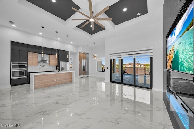 kitchen featuring ceiling fan, wall chimney exhaust hood, decorative light fixtures, a kitchen island with sink, and appliances with stainless steel finishes