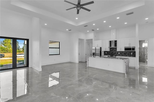 kitchen with a towering ceiling, stainless steel appliances, a tray ceiling, a kitchen island with sink, and white cabinets