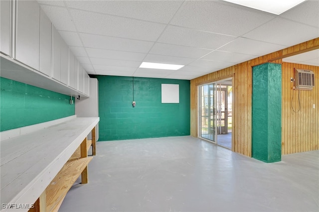 basement featuring a wall mounted air conditioner, a drop ceiling, and wooden walls