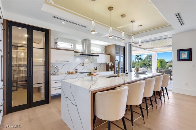 kitchen featuring a center island with sink, a tray ceiling, decorative light fixtures, island range hood, and white cabinetry