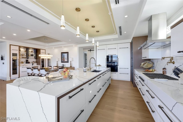 kitchen with wall chimney exhaust hood, hanging light fixtures, a spacious island, a tray ceiling, and white cabinets