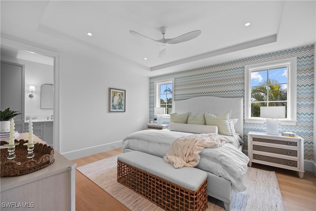 bedroom featuring light wood-type flooring, ceiling fan, ensuite bathroom, and a tray ceiling