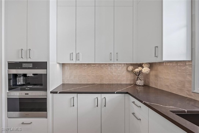 kitchen featuring white cabinets, backsplash, stainless steel double oven, and dark stone counters