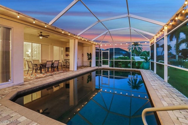 pool at dusk featuring glass enclosure, ceiling fan, and a patio area