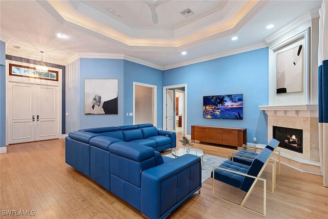 living room with ceiling fan with notable chandelier, a tray ceiling, crown molding, a premium fireplace, and light hardwood / wood-style floors