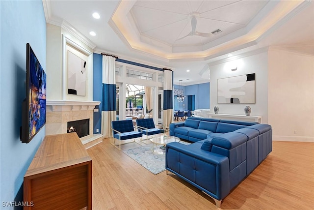 living room featuring a raised ceiling, a tiled fireplace, crown molding, and light hardwood / wood-style flooring