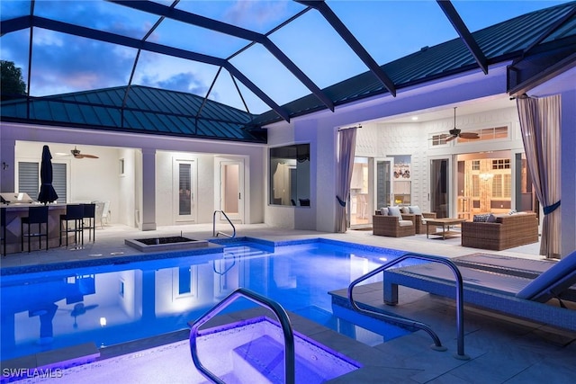 pool at dusk featuring glass enclosure, an outdoor living space, ceiling fan, and exterior bar