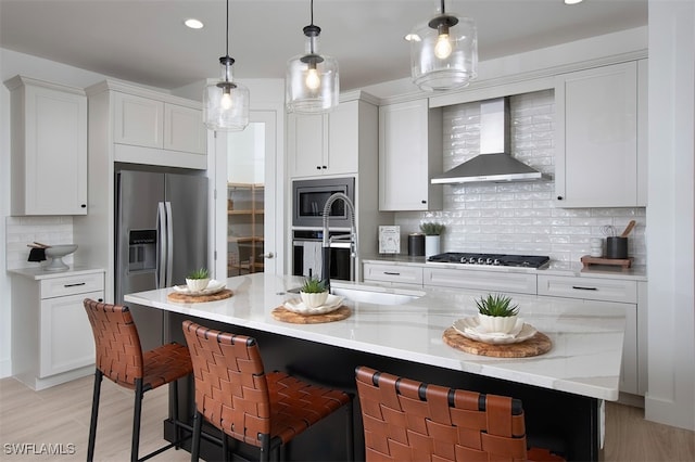 kitchen with white cabinets, wall chimney range hood, hanging light fixtures, stainless steel appliances, and a center island with sink