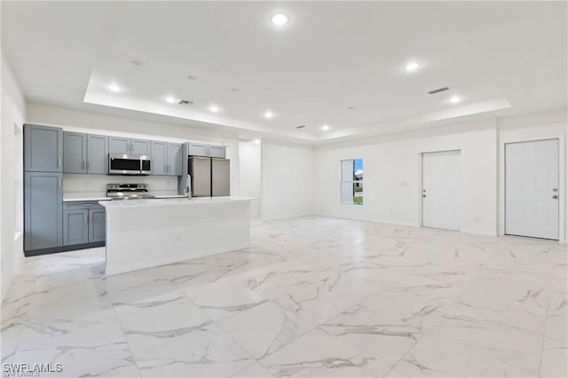 kitchen with appliances with stainless steel finishes, a raised ceiling, a kitchen island with sink, and gray cabinetry