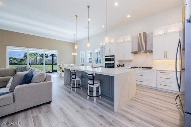 kitchen with white cabinetry, hanging light fixtures, wall chimney range hood, light hardwood / wood-style flooring, and a kitchen island with sink